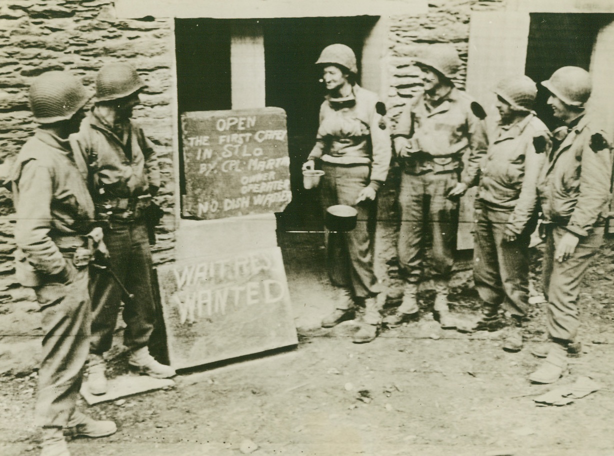 Man Shortage There, Too, 7/21/1944. St. Lo, France – Prominently displayed beneath the sign advertising the opening on the first canteen is a placard seeking waiters. Cpl. E. L. Martin, Martinsville, Virginia, stands at the head of the line with his canteen cup and pan, waiting for the mess call. Credit: U.S. Army Radiotelephoto from ACME;