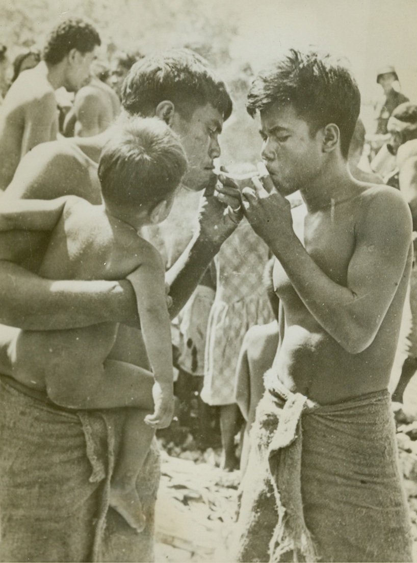 Lighting Up, 1/7/1944. Butaritari Island, Makin – Holding his naked son, a Makin native is about to enjoy the luxury of an American cigarette as he grubs a light from one of his neighbors.  This crowd of natives, huddled on rocky Red beach, is about to be evacuated from the area by Americans who hold the beach. Credit line (U.S. Signal Corps photo from ACME);
