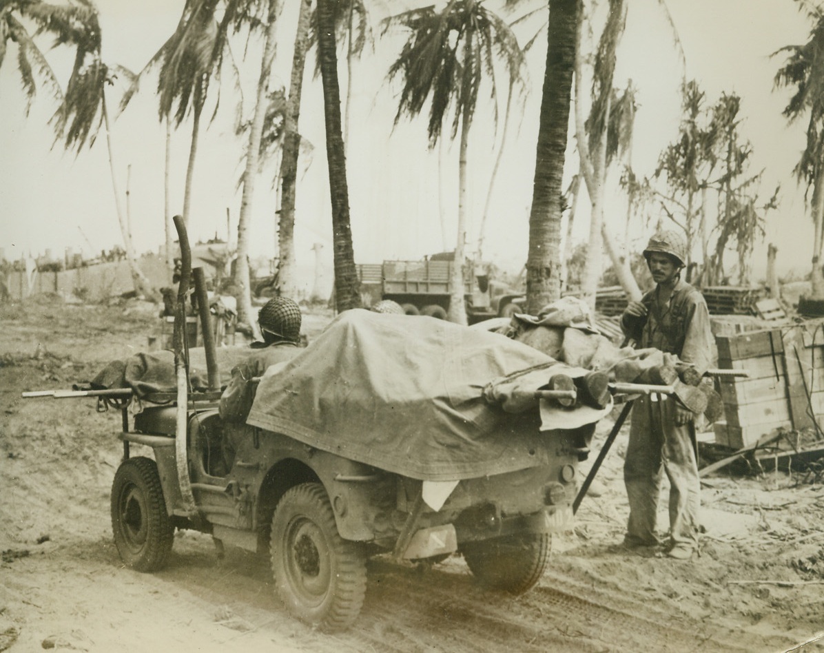JEEP SERVES AS A HEARSE, TOO, 3/6/1944. ENIWETOK ISLAND—The versatile Jeep takes on a grim task on Eniwetok Island. Covered by a tarpaulin, bodies of American dead are carried away for burial on the island. Soldier at right sadly watches as his buddies take their last ride. Credit Line (ACME);