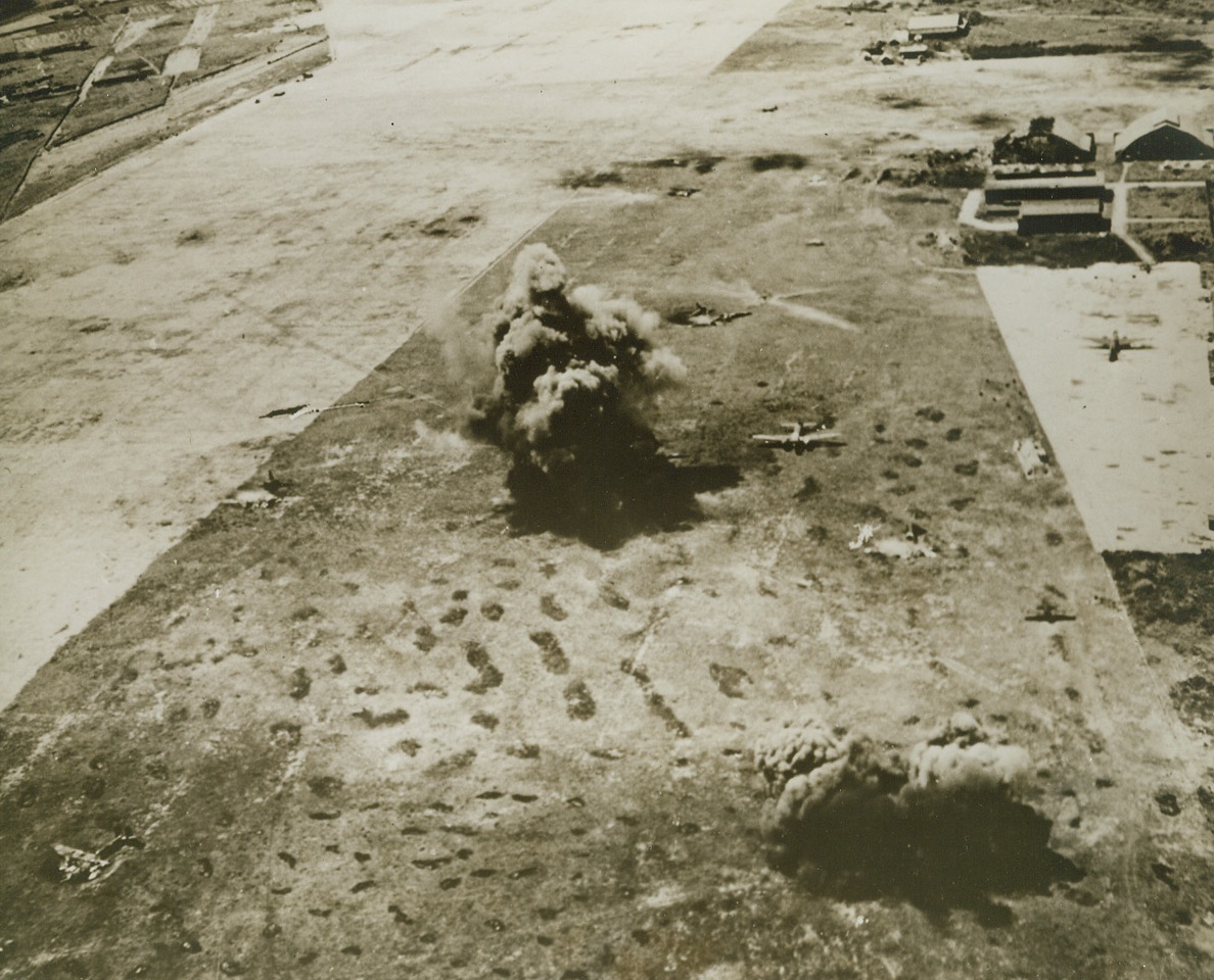 Japs Grounded—But Good. Huge billows of black smoke and dirt mushroom above the runway at the Japanese base at Saipan in the Marianas, as U.S. Navy carrier based planes blasted the field and installations last February 23rd. Nip planes which were caught “Flat-Footed” dot the runway—some of them already wrecked by the attackers. Credit: U.S. Navy Photo from Acme;