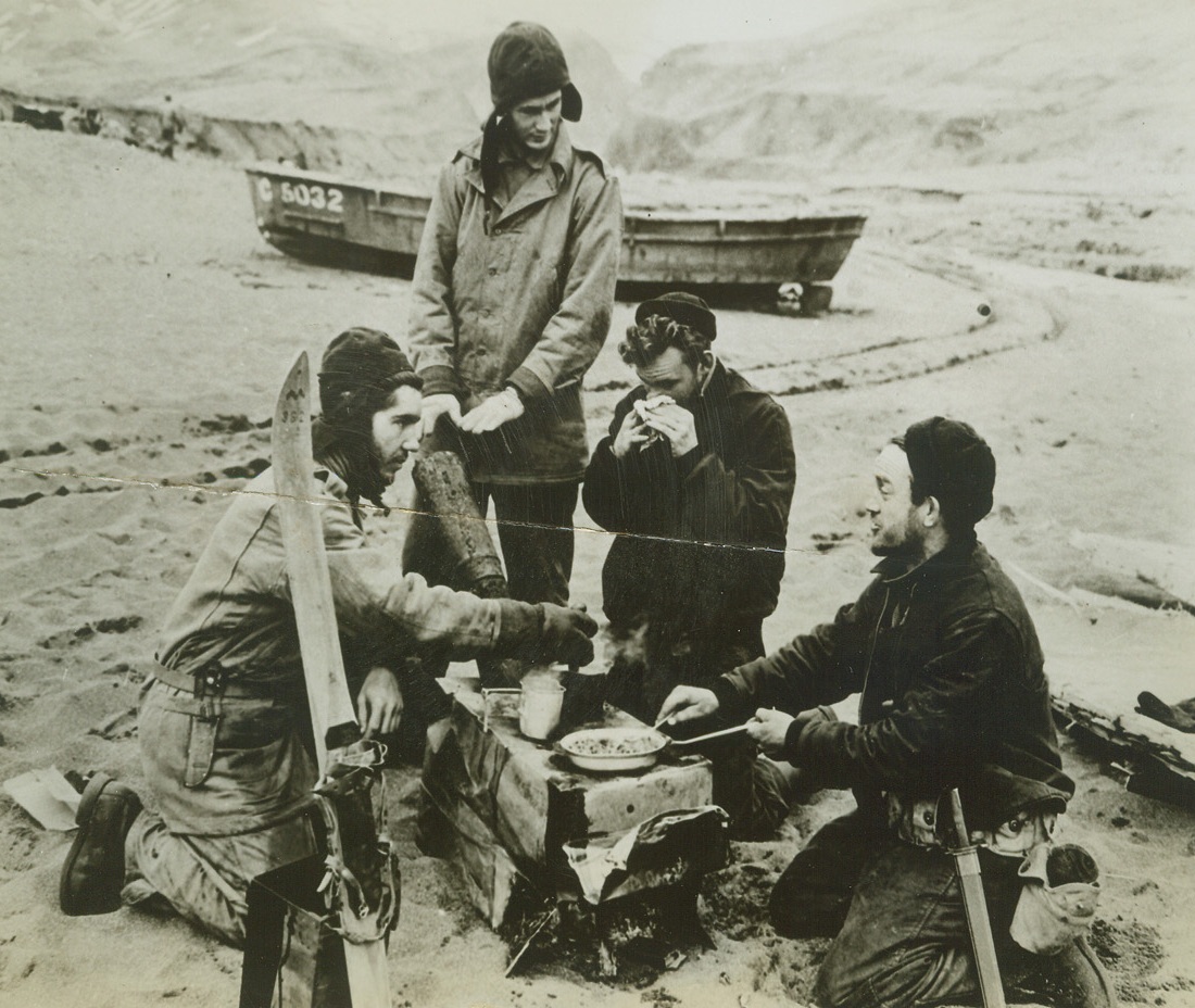 Double Purpose, 5/29/1943. Attu – A warm fire on Attu’s chilly beach serves a double purpose.  It cooks a hot meal and thaws out the hands of these U.S. sailors in the Holtz Bay area of Attu island, following the landing there to drive out or destroy the Japanese Garrison.  Even in May that cold-weather garb and those skies (foreground) come in handy in the Aleutians.Credit (Official US Navy photo from ACME);