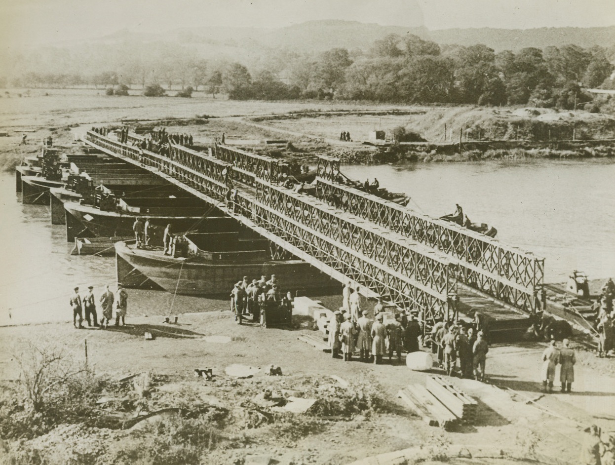Enemy Demolition Go Hang, 6/29/1944. England—A Bailey Bridge, new U.S.-British war creation proving of immense value to Allied operations and an answer to enemy demolition, is shown being constructed across a river in England. Parts of the bridge, built both in the U.S. and England, are interchangeable and can be speedily put together. It was invented by Mr. D.C. Bailey of the Ministry of Supply. Credit: British official photo from ACME.;