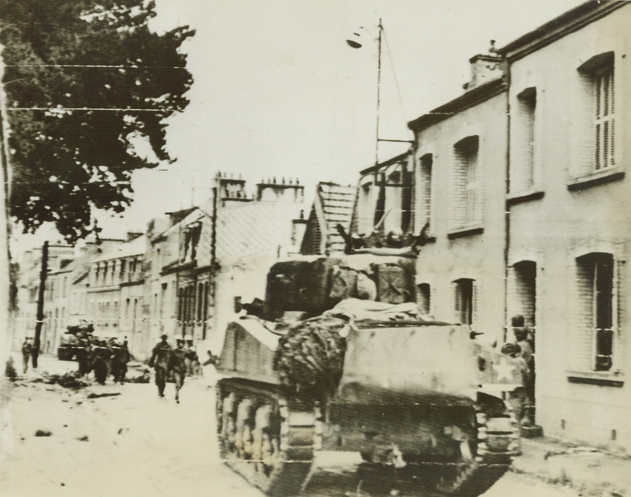 Yanks Make Tank Tracks In Cherbourg, 6/27/1944. Cherbourg, France – While Yank soldiers seek shelter in doorways from German snipers, American tanks move through the streets of Cherbourg immediately before the fall of the port city was officially announced. Now that American forces are in possession of Cherbourg, the Allies have at their disposal an adequate port for the landing of reinforcements and supplies. Credit: Army Radiotelephoto from ACME;