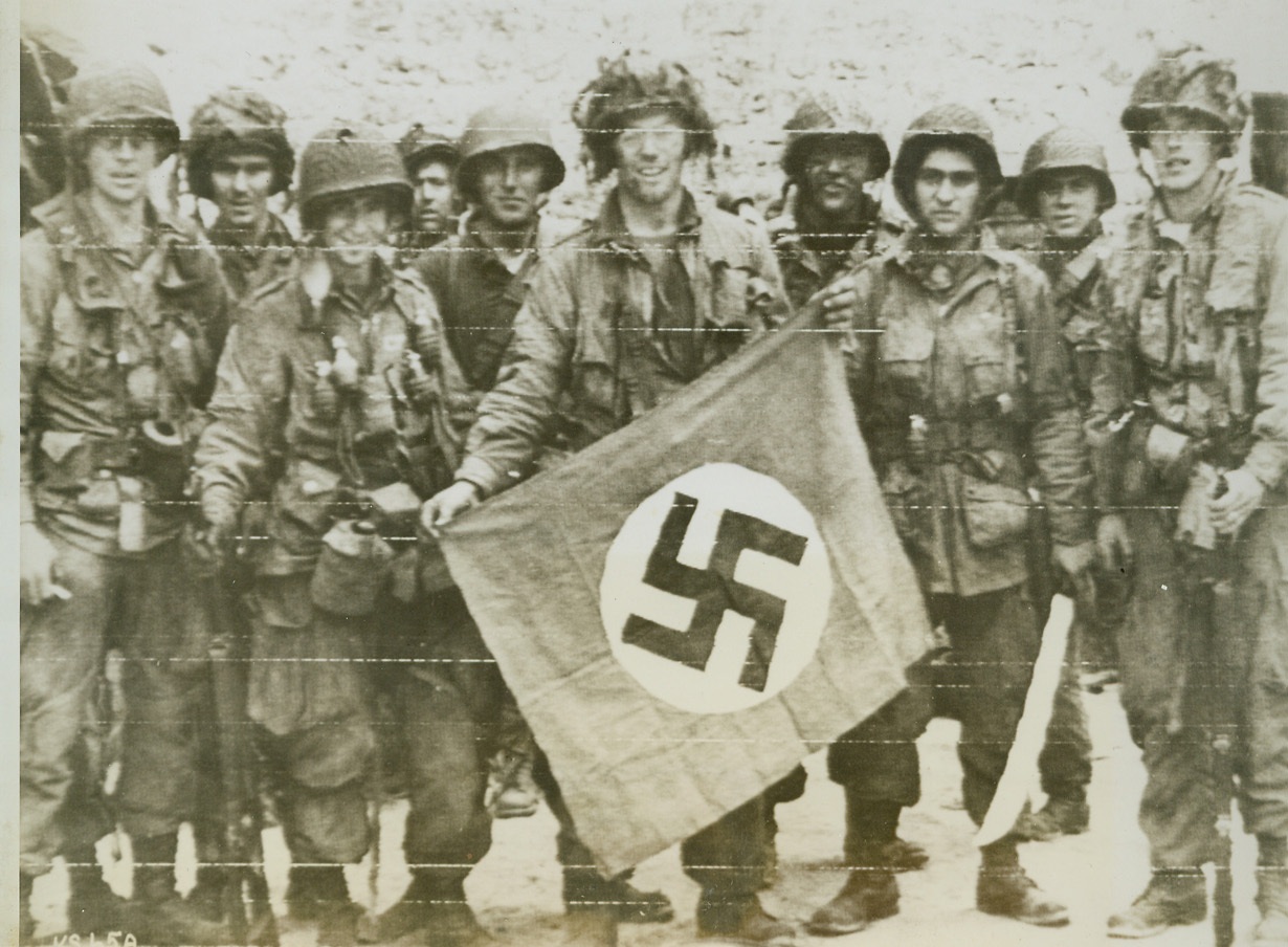 First Booty Brings Cheer, 6/9/1944. France—American paratroopers smile happily as they display a captured Nazi flag taken in assault on a French village. These soldiers were among the first Allies to return to France since the retreat from Dunkirk. Credit: Signal Corps radiotelephoto from ACME;
